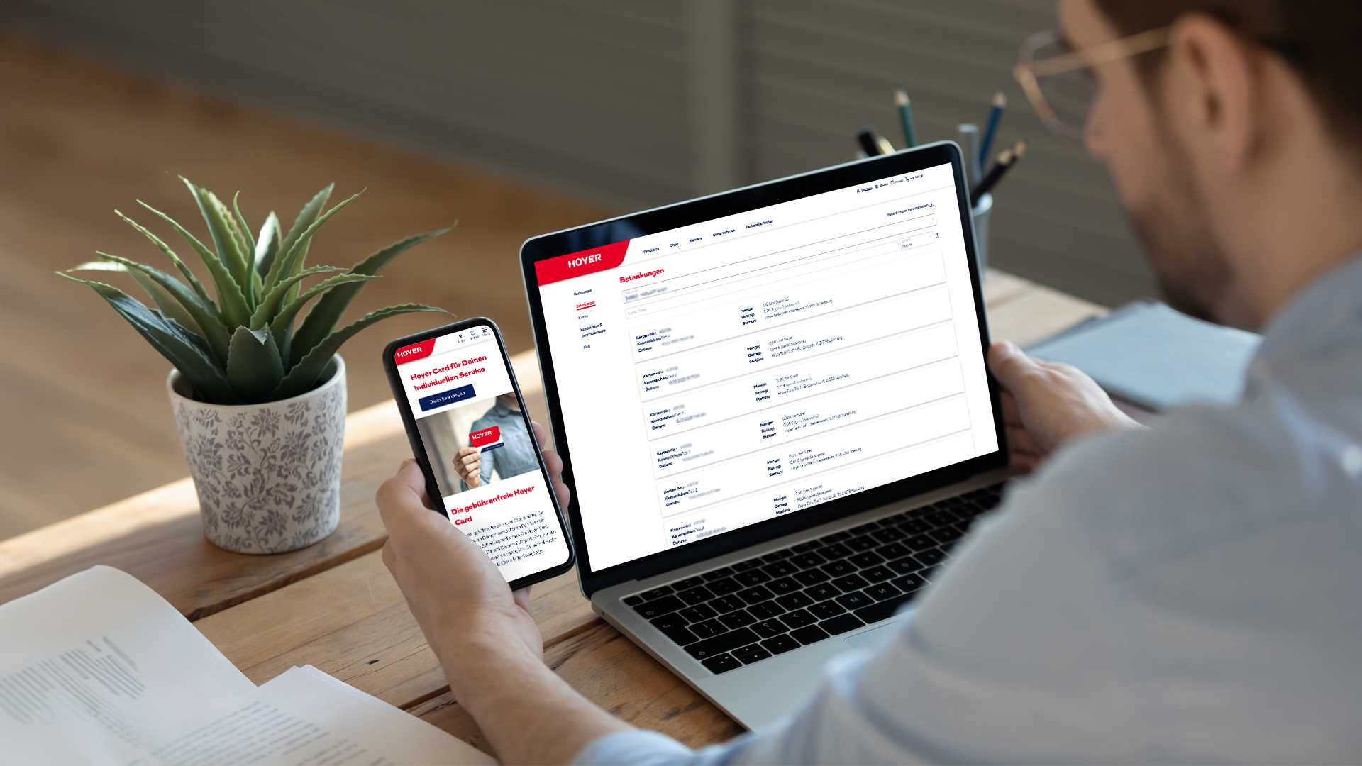 Blick über Schulter eines Mannes, der vor einem Laptop sitzt und gleichzeitig ein Smartphone in der Hand hat
