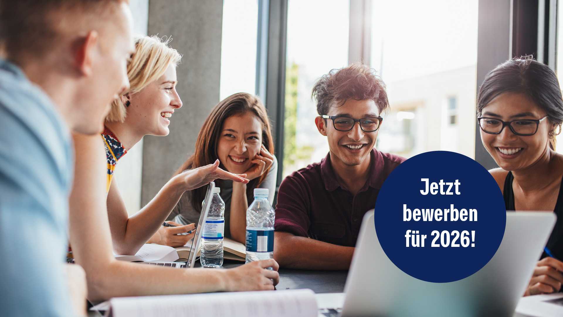Fünf Menschen sitzen an einem Tisch im Halbkreis. Einige schauen gemeinsam auf einen Laptop-Bildschirm