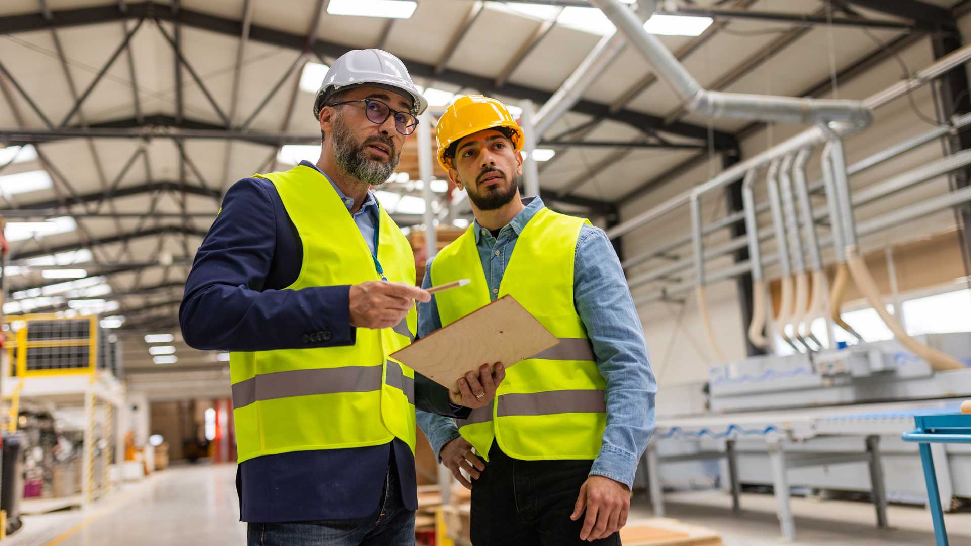 Zwei Menschen mit gelben Warnwesten und Helmen stehen in einer Produktionshalle